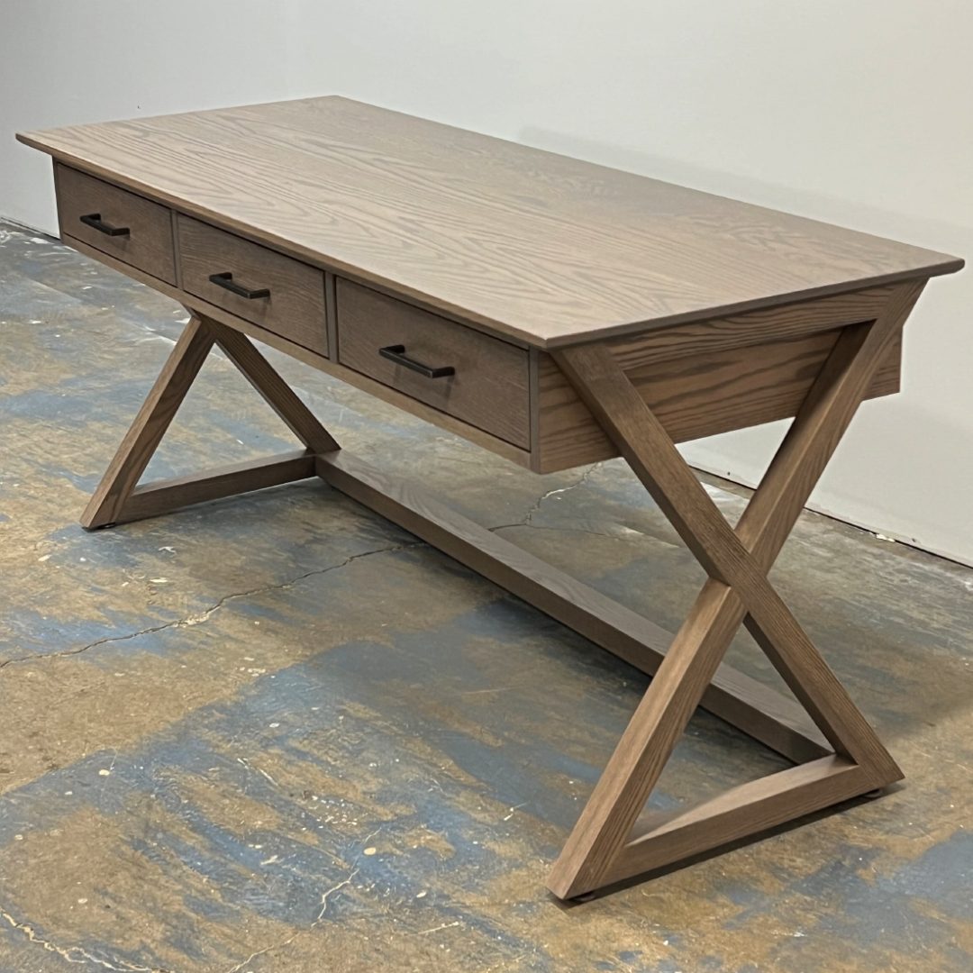 A Modern Writing Desk With A Grey-Brown Wood Finish, Viewed From The Side. The Desk Stands On Unique X-Shaped Wooden Legs And Features A Flat Tabletop With Three Slender Drawers Across The Front For Storage.